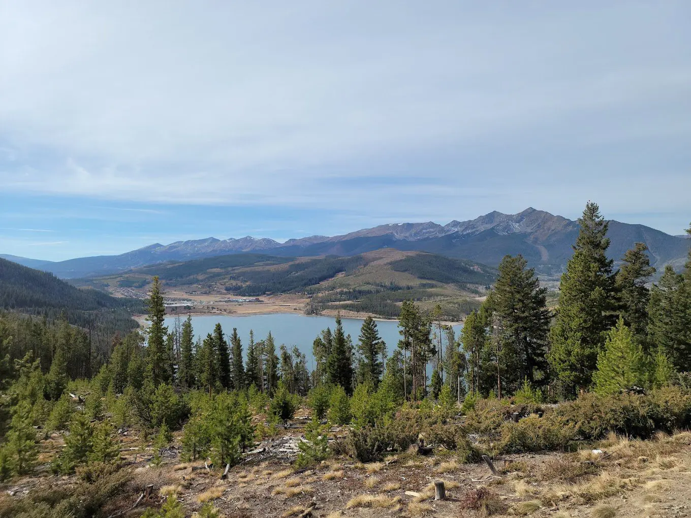 anoramic view of a mountain lake, which is one of the top things to do in Silverthorne CO, as seen from an overlook. The image features a blue lake surrounded by forested hills and large, dark mountains in the distance under a partly cloudy sky. Pine trees frame the view in the foreground."